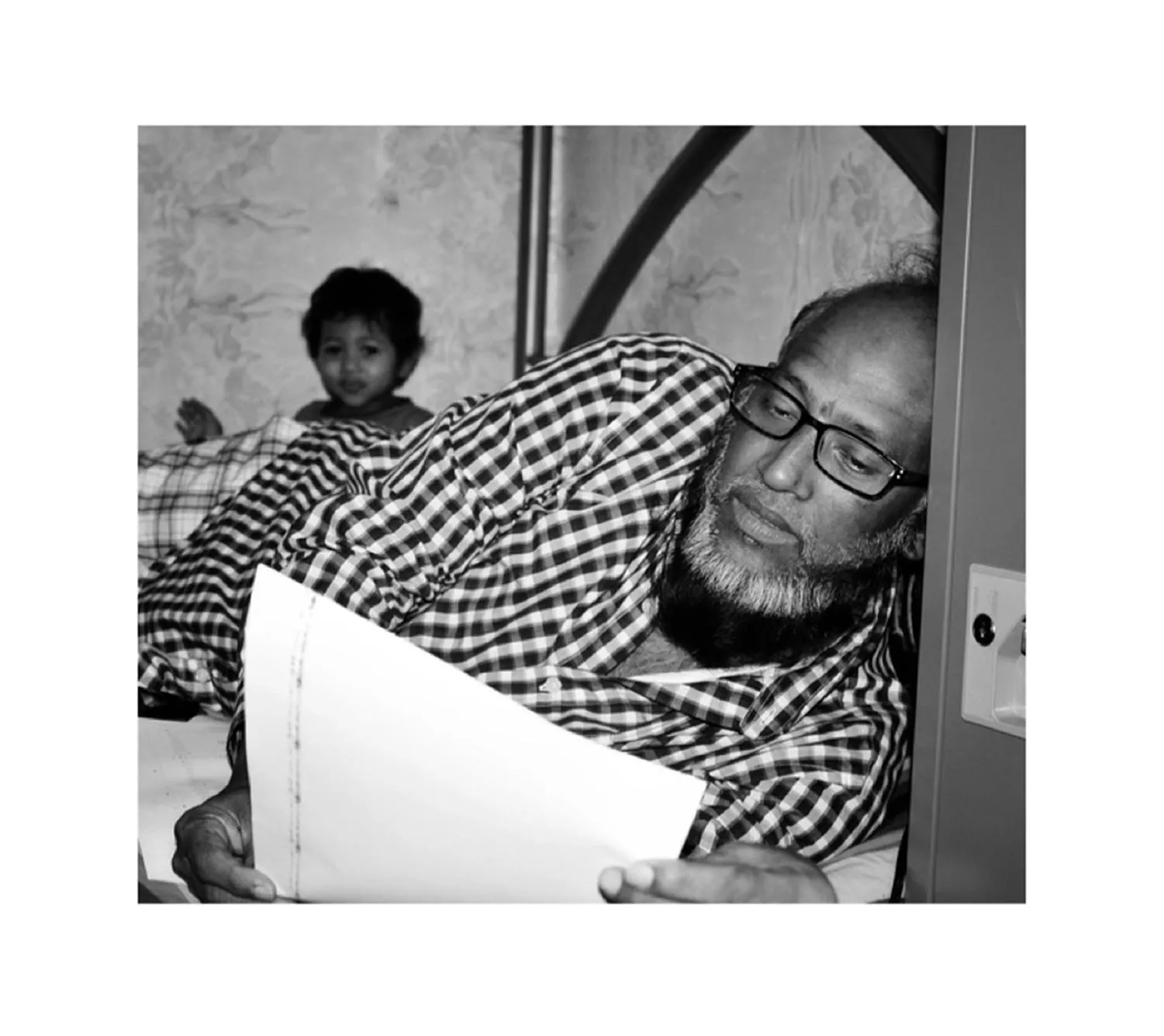 Black and white image of a man laying on a bed, reading a sheet of paper, while a boy looks on in the background. 